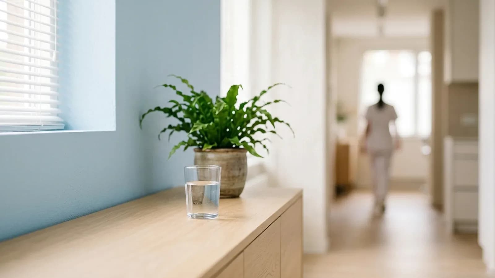 Ruhige Ecke eines hellen Praxisraums mit Wasserglas und Grünpflanze vor pastellblauer Wand, dahinter ein unscharfer Flur
